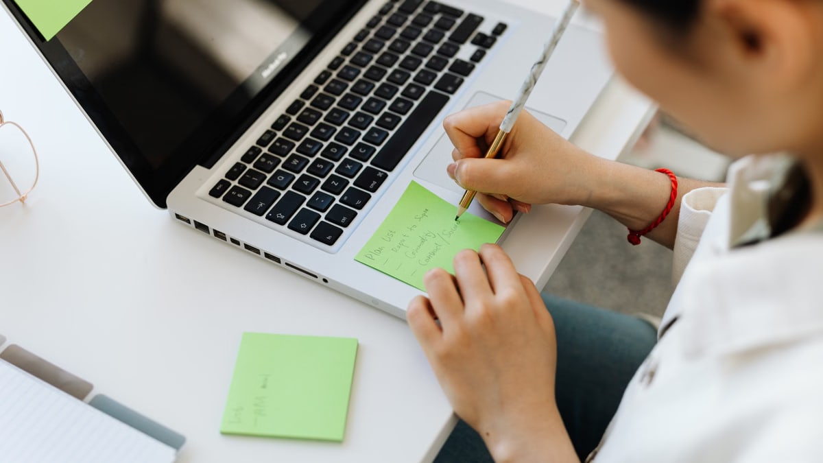 Person writing a to-do list on sticky note next to laptop for organization.